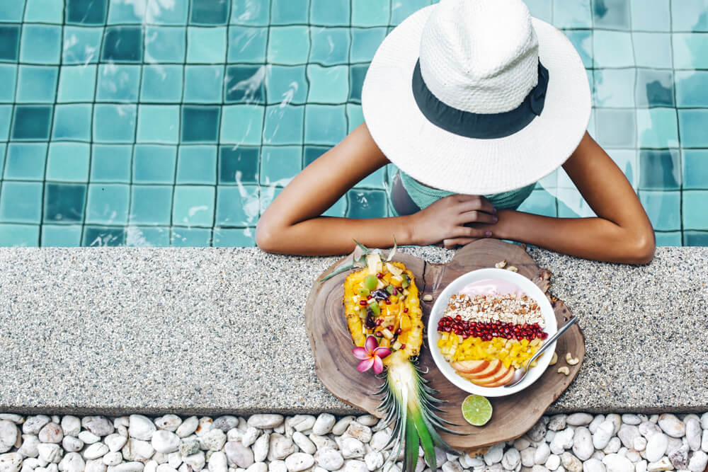 Žena s bílým kloboukem u okraje bazénu se smoothie bowl a ananasem